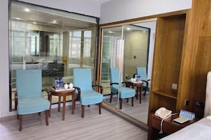 Habitación de hotel con un espejo grande, dos sillones azules, una mesita con un teléfono y una libreta, y un armario de madera.