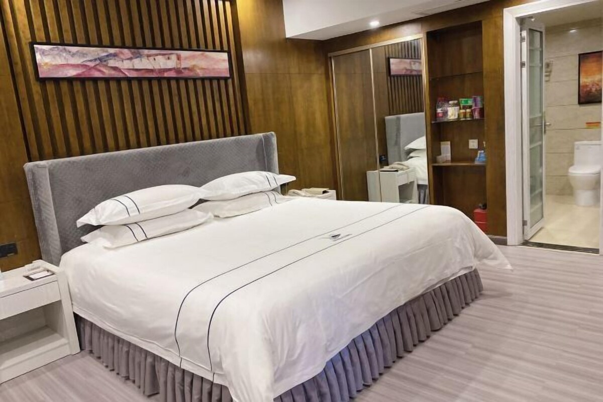 A modern hotel room with a large bed, a bedside table, a wardrobe, and a bathroom visible through an open door.