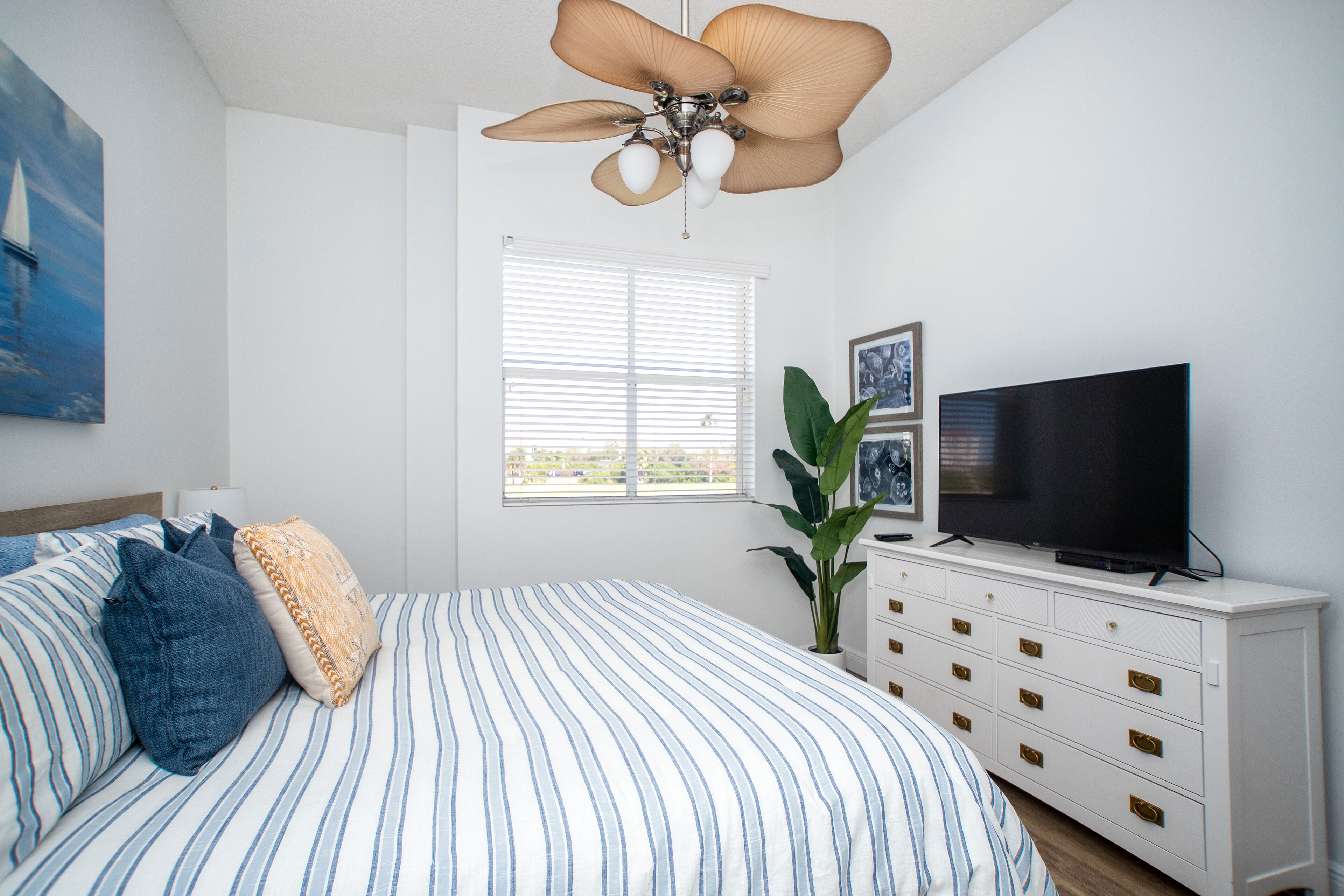 Un dormitorio con cama, televisor, cómoda, ventilador de techo, ventana y planta.