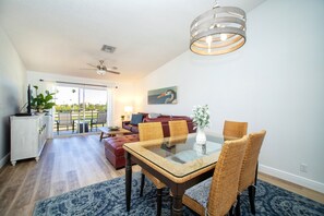 Un salon spacieux avec une table à manger au plateau en verre, des chaises en osier, un ventilateur de plafond et une grande fenêtre donnant sur l’extérieur.