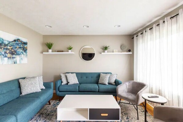 Un salon moderne avec un canapé de couleur turquoise, une table basse blanche et un fauteuil gris. On y trouve deux étagères blanches ornées de plantes et un miroir rond placé au-dessus.