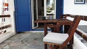 Une table et des chaises en bois sont disposées sur un balcon, devant une porte bleue et sur un fond de murs blancs.