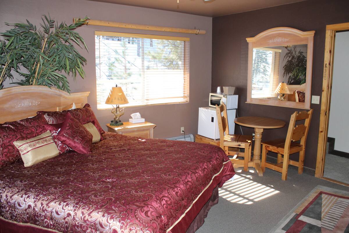 A bedroom with a large bed, a desk, a chair, a small refrigerator, a lamp, and a mirror.