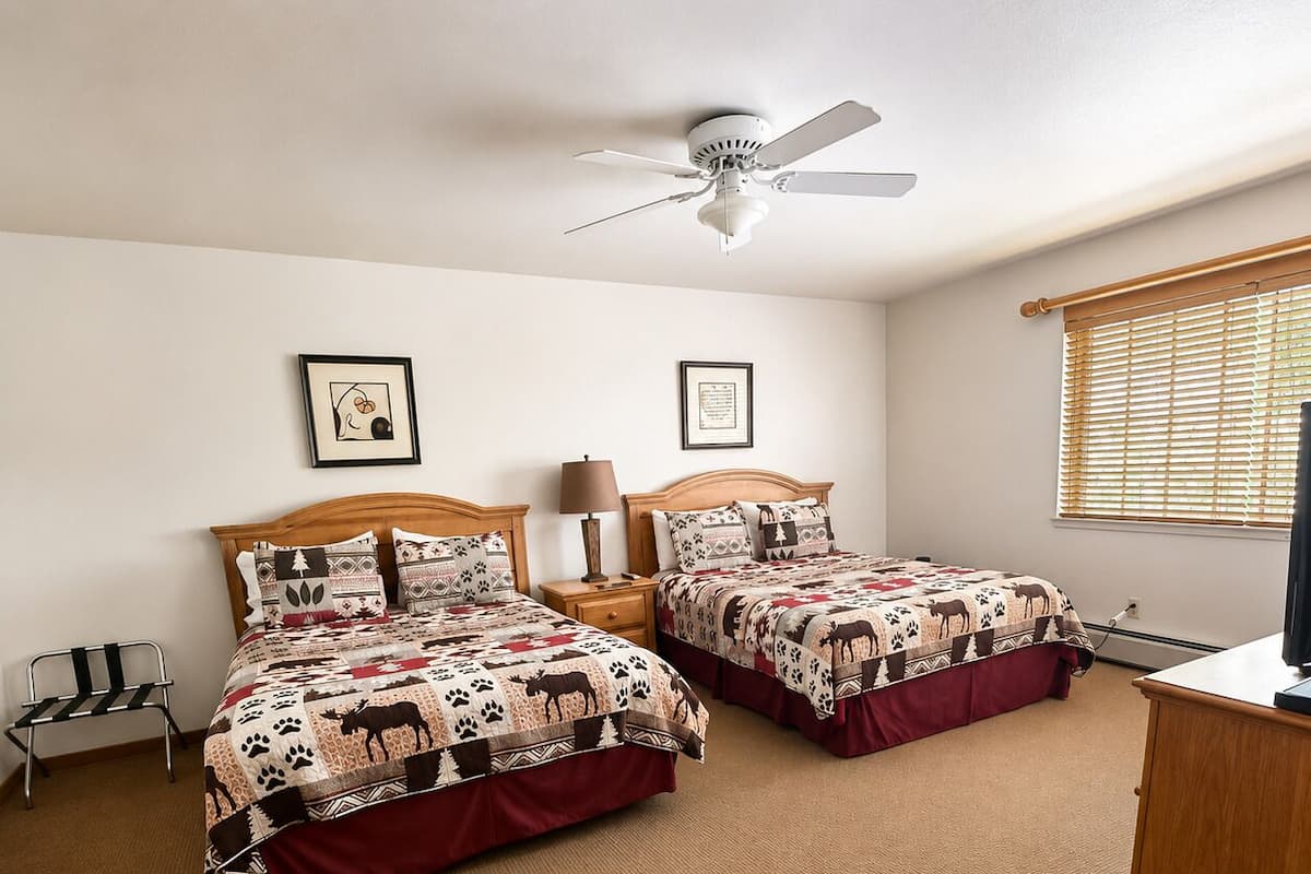 A room with two beds, a ceiling fan, a window with blinds, a desk, and a trash can.