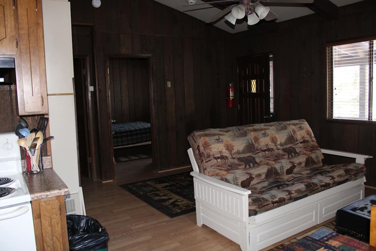 A room with a white sofa, wooden walls, a ceiling fan, a window with blinds, and a kitchen area with a stove and utensils.