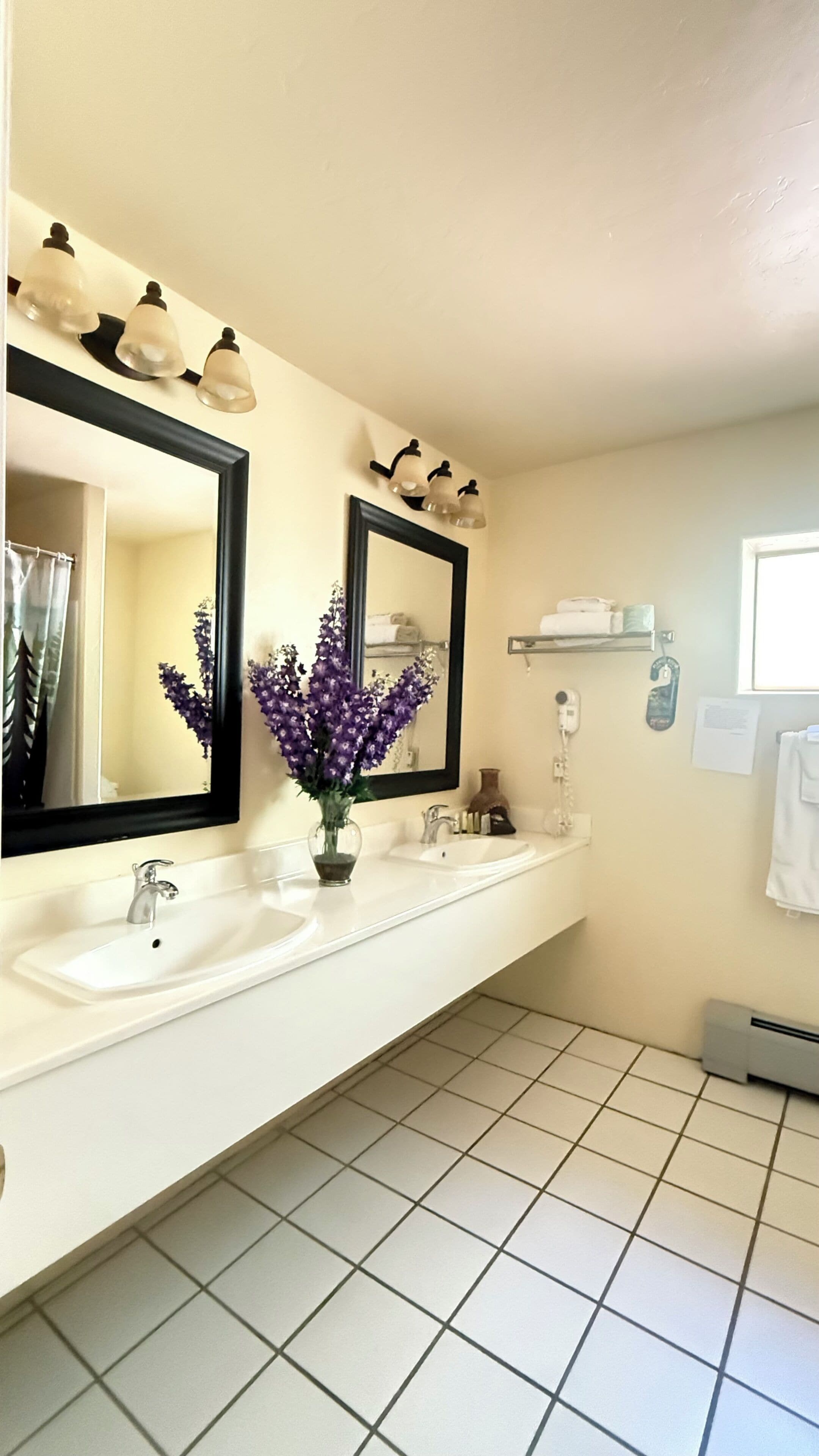 Ein Badezimmer mit einem weißen Waschbecken, zwei Spiegeln, einer Vase mit violetten Blumen und einer Duschvorhang.