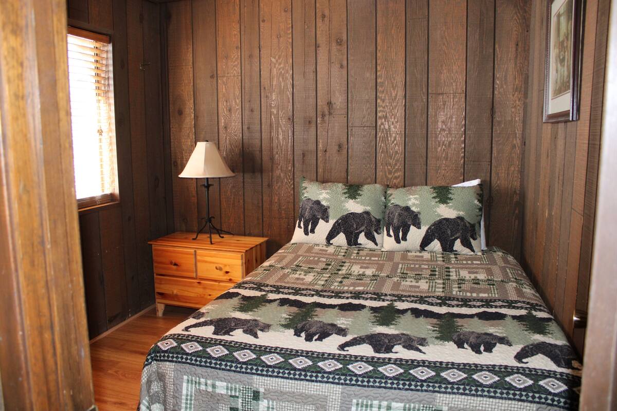 A bedroom with a wooden wall, a bed with a bear-themed bedspread, a nightstand with a lamp, and a window with blinds.