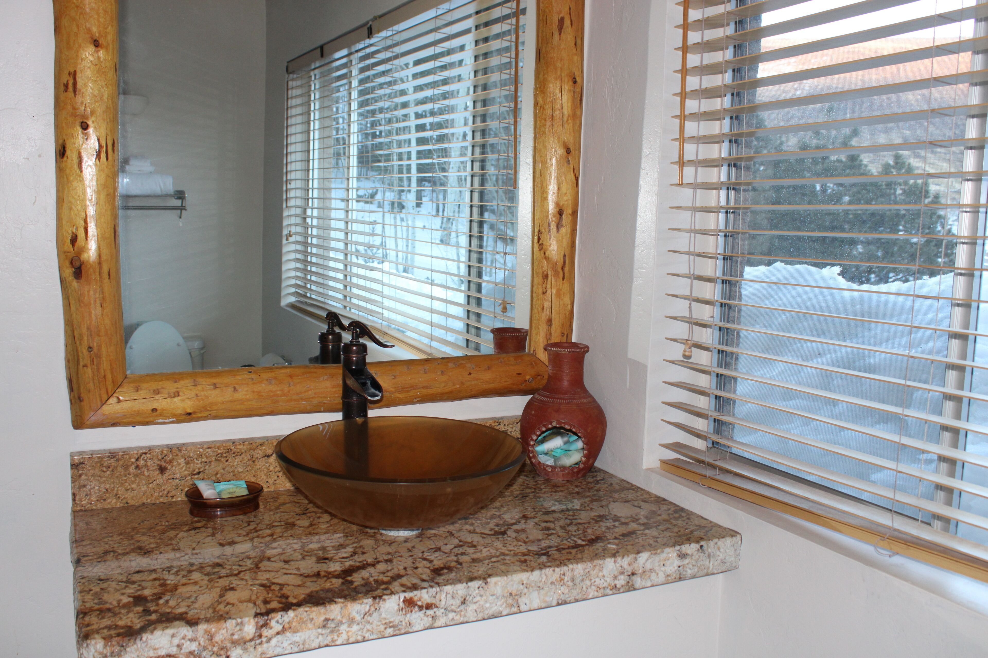 A bathroom with a wooden-framed mirror, a glass bowl sink, and a red vase on a granite countertop.