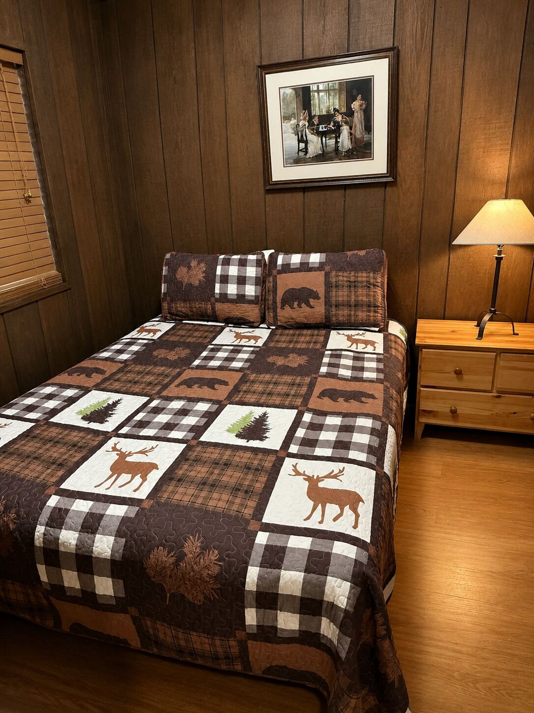 A bedroom with a wooden wall, a framed picture, a bedside table with a lamp, and a quilt with animal prints.