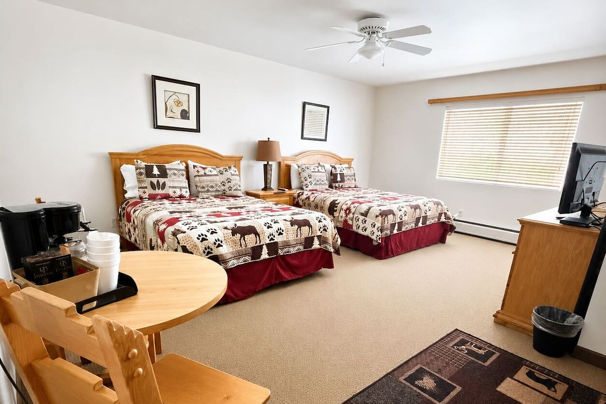 A room with two beds, a desk with a chair, a ceiling fan, a window with blinds, and a television.