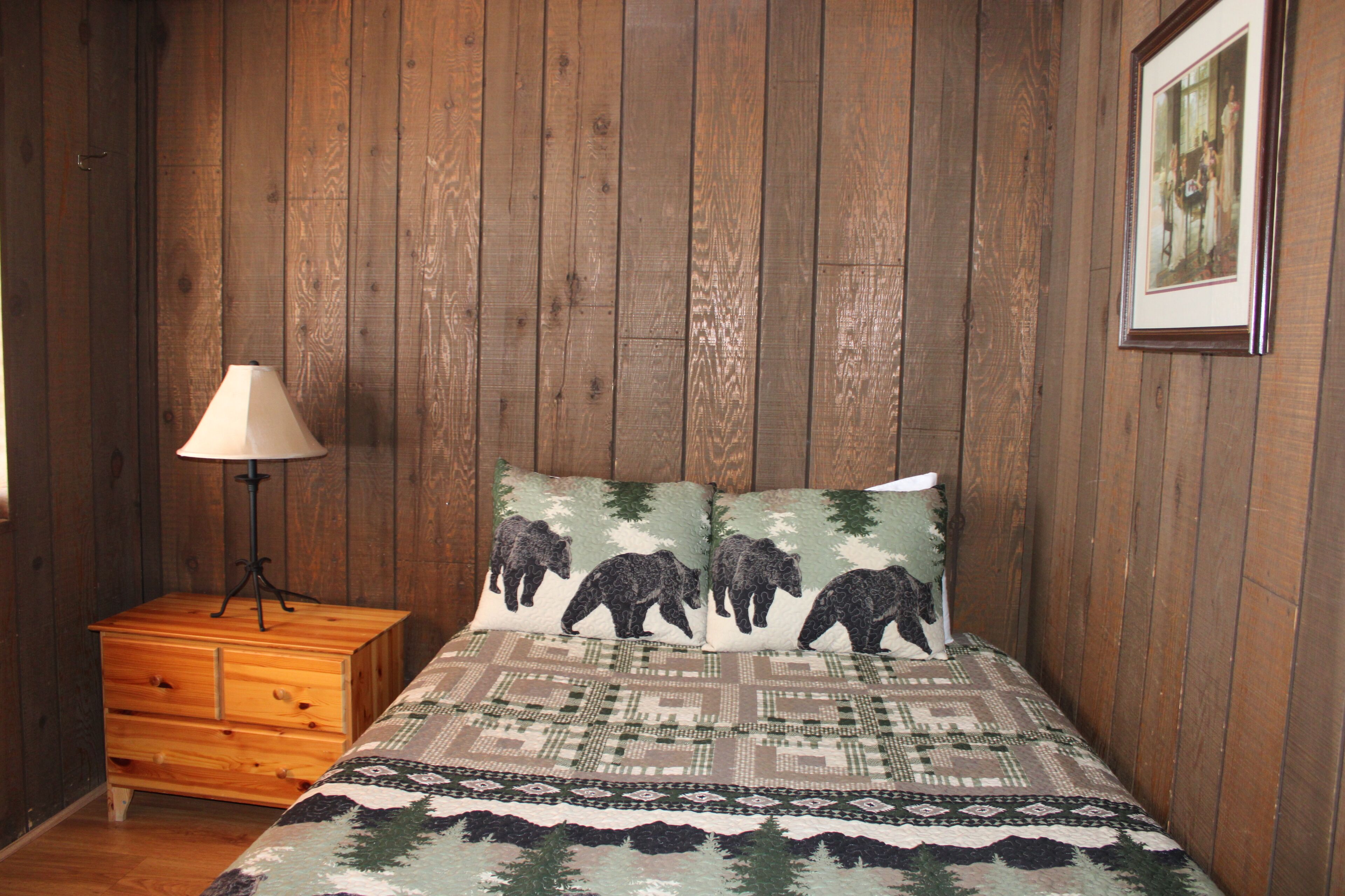 Une chambre avec des murs recouverts de lambris en bois, une table de chevet en bois avec une lampe, et un lit avec une couverture à motifs et deux oreillers décorés de motifs d’ours.