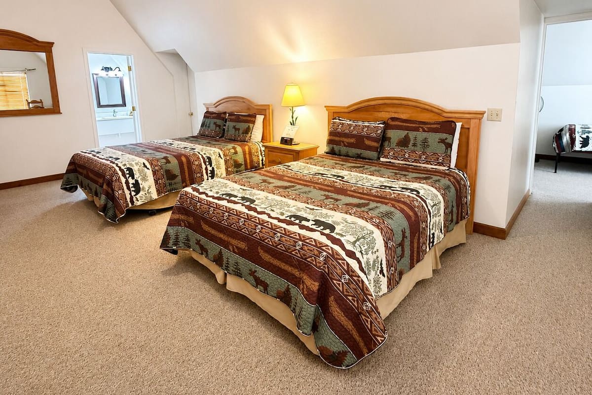 A bedroom with two beds, a nightstand with a lamp, a mirror, and a door leading to a bathroom.
