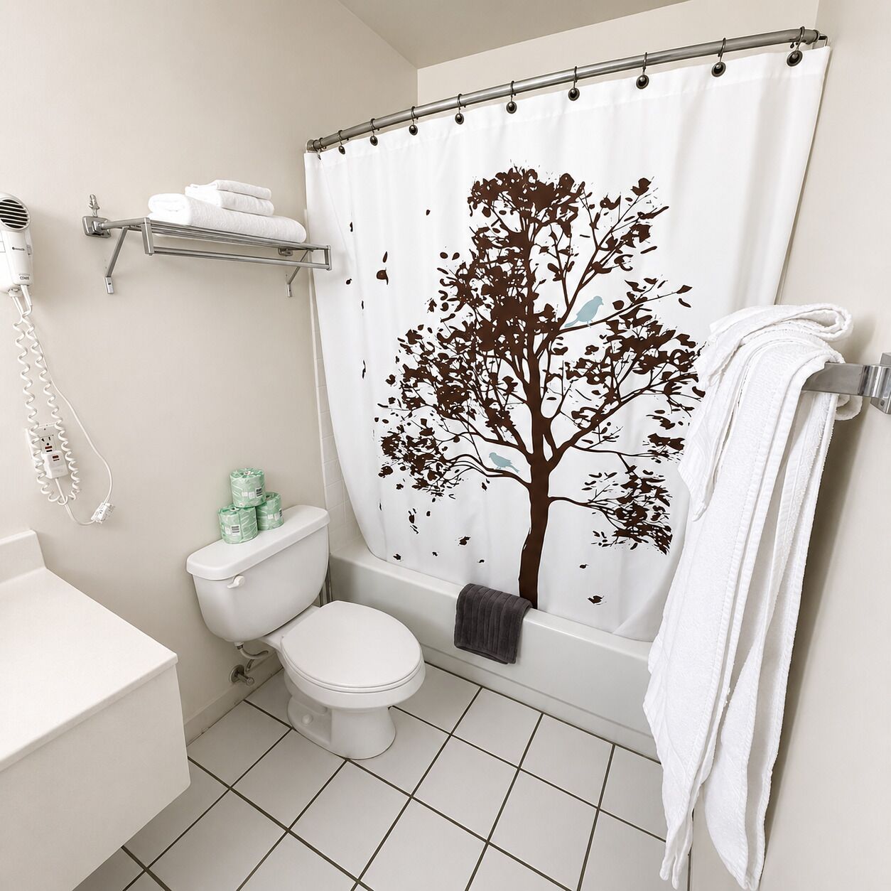 Une salle de bain avec des toilettes blanches, une baignoire dotée d’un rideau de douche orné d’un motif d’arbre, et un porte-serviettes avec une serviette.