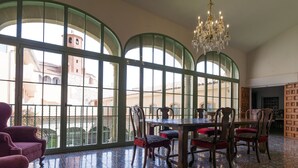 Un comedor con una gran mesa y sillas, ventanas arqueadas amplias, una araña y baldosas de suelo estampadas.
