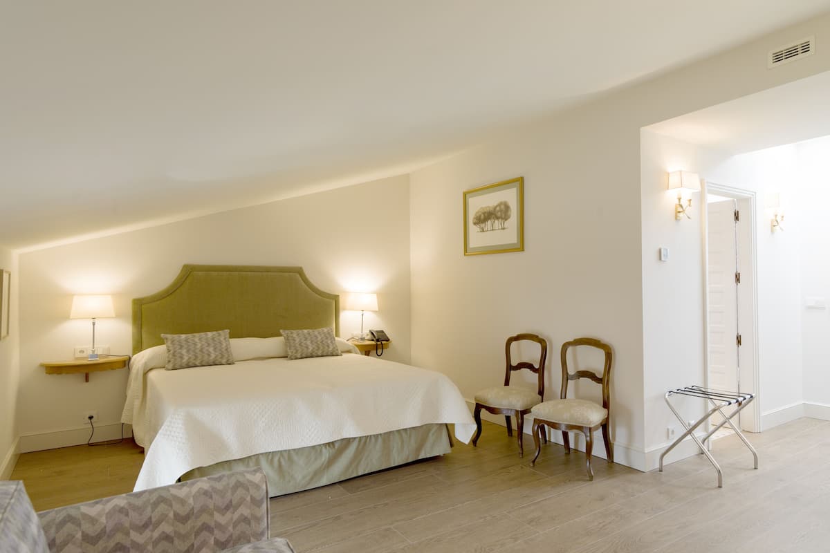 Quarto de hotel com uma cama grande, duas mesas de cabeceira com luminárias, duas cadeiras, um quadro na parede e uma porta que dá para outro cômodo.