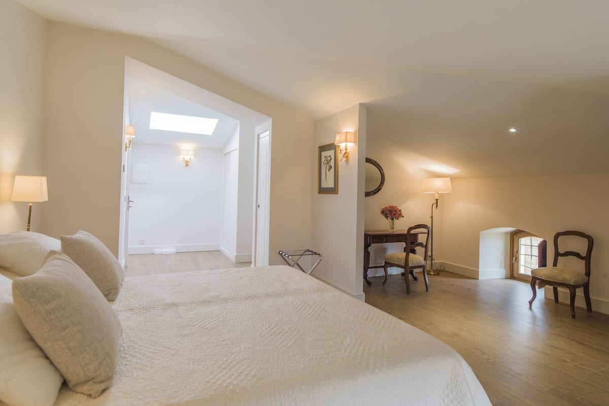 Quarto com uma cama grande, uma mesa e uma cadeira. Há uma porta que dá para outro cômodo.