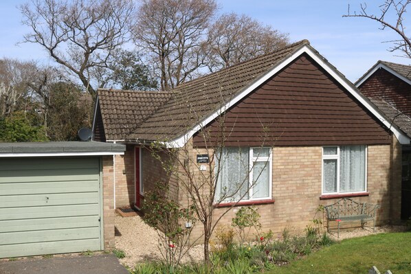 A single-story house with a brown roof, a brick facade, and a small front yard with a bench and a tree.