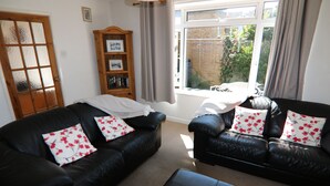 A living room with two black leather sofas, each with floral cushions, a wooden bookshelf, and a large window with curtains.