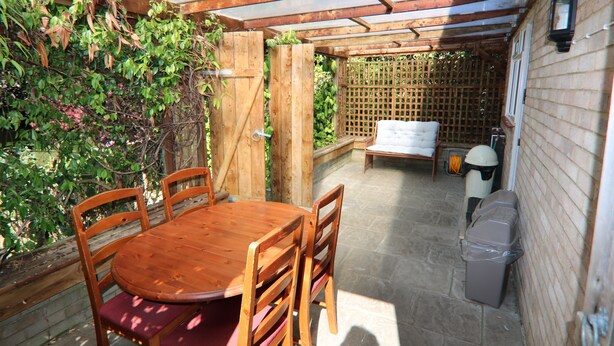 A wooden table and chairs under a glass roof, surrounded by a wooden fence and greenery.