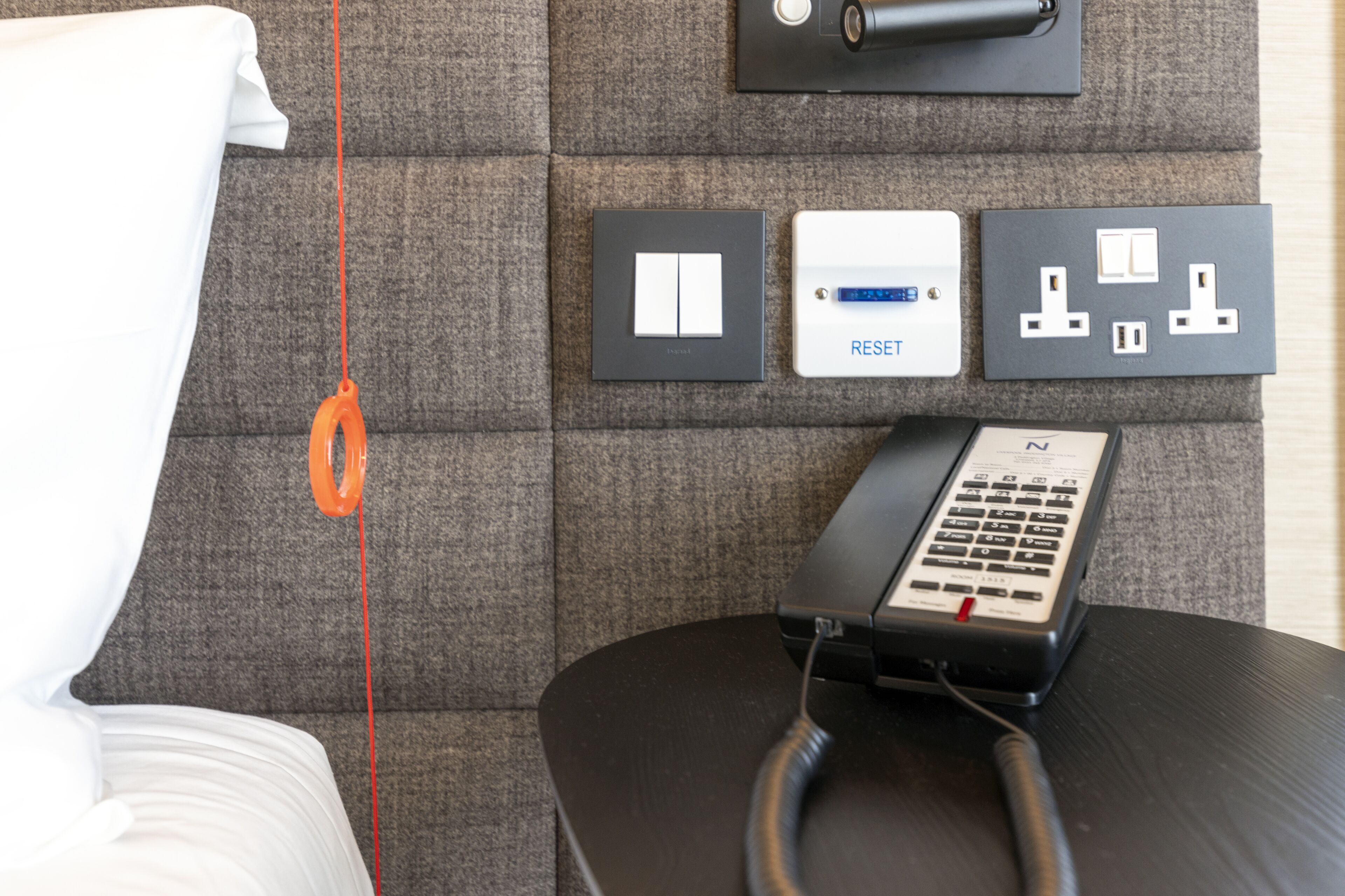 Habitación de hotel con una cama, un panel de control con un cable rojo, un tablero de interruptores en la pared y un teléfono.
