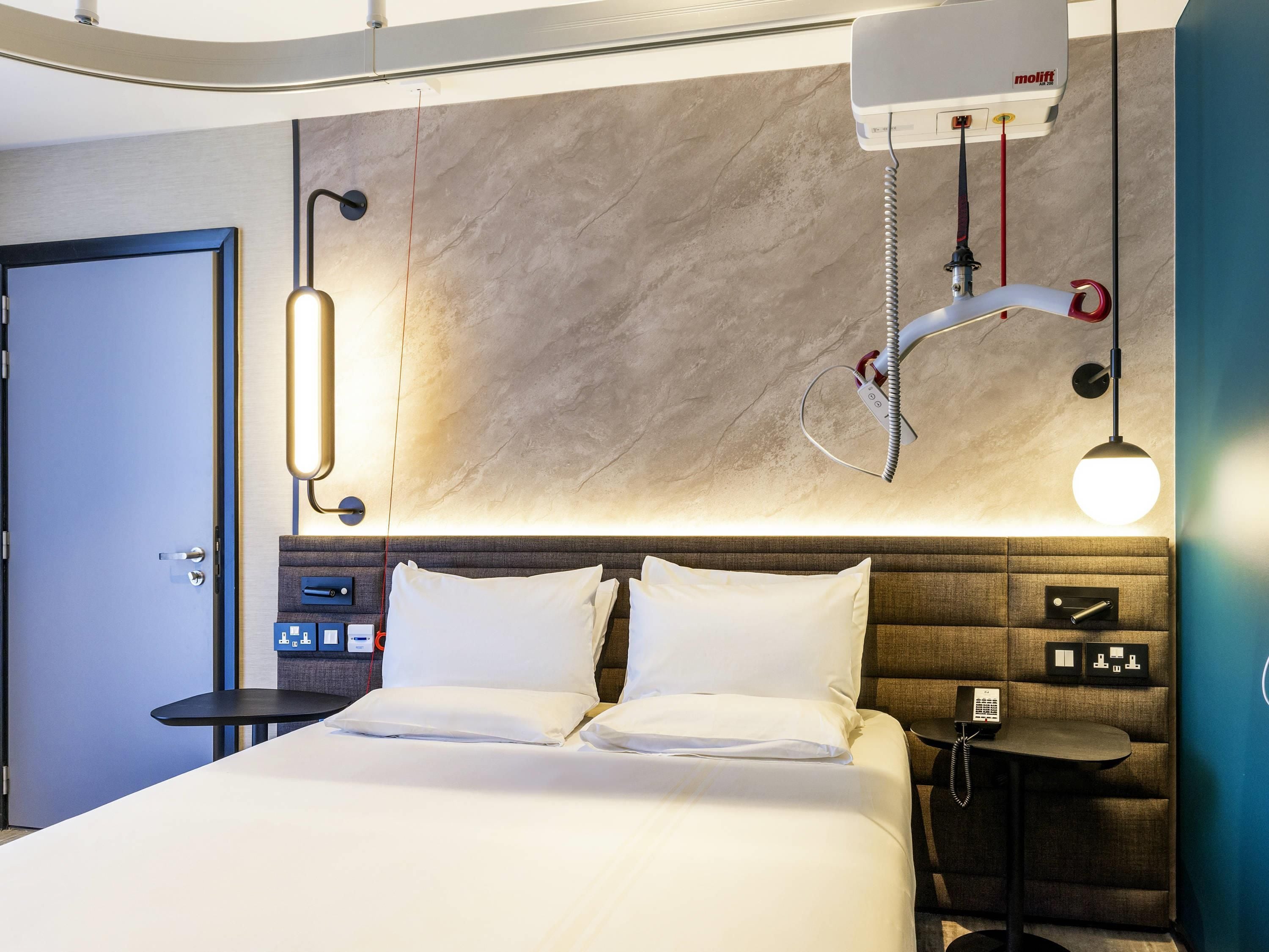 A hotel room with a bed, bedside table, lamp, and a wall-mounted air conditioning unit.