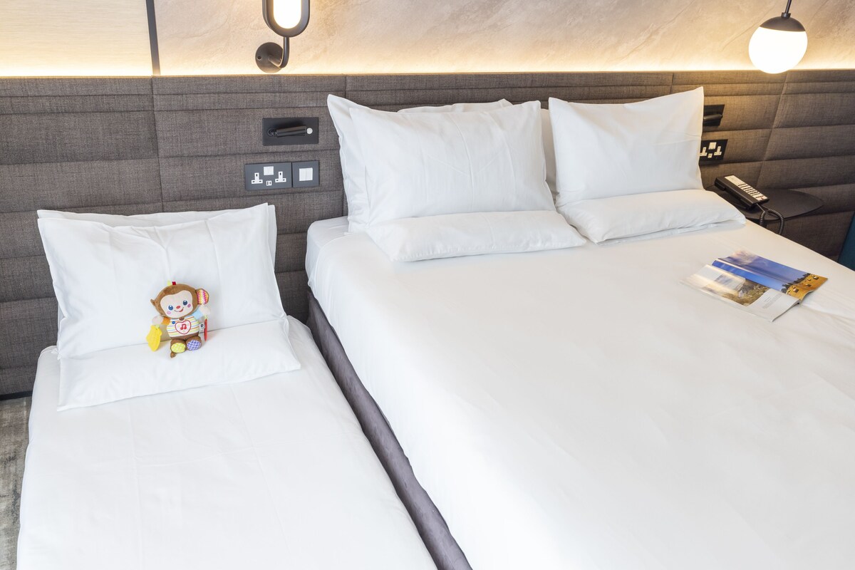 A hotel room with two beds, a nightstand with a phone and lamp, and a plush toy on one bed.
