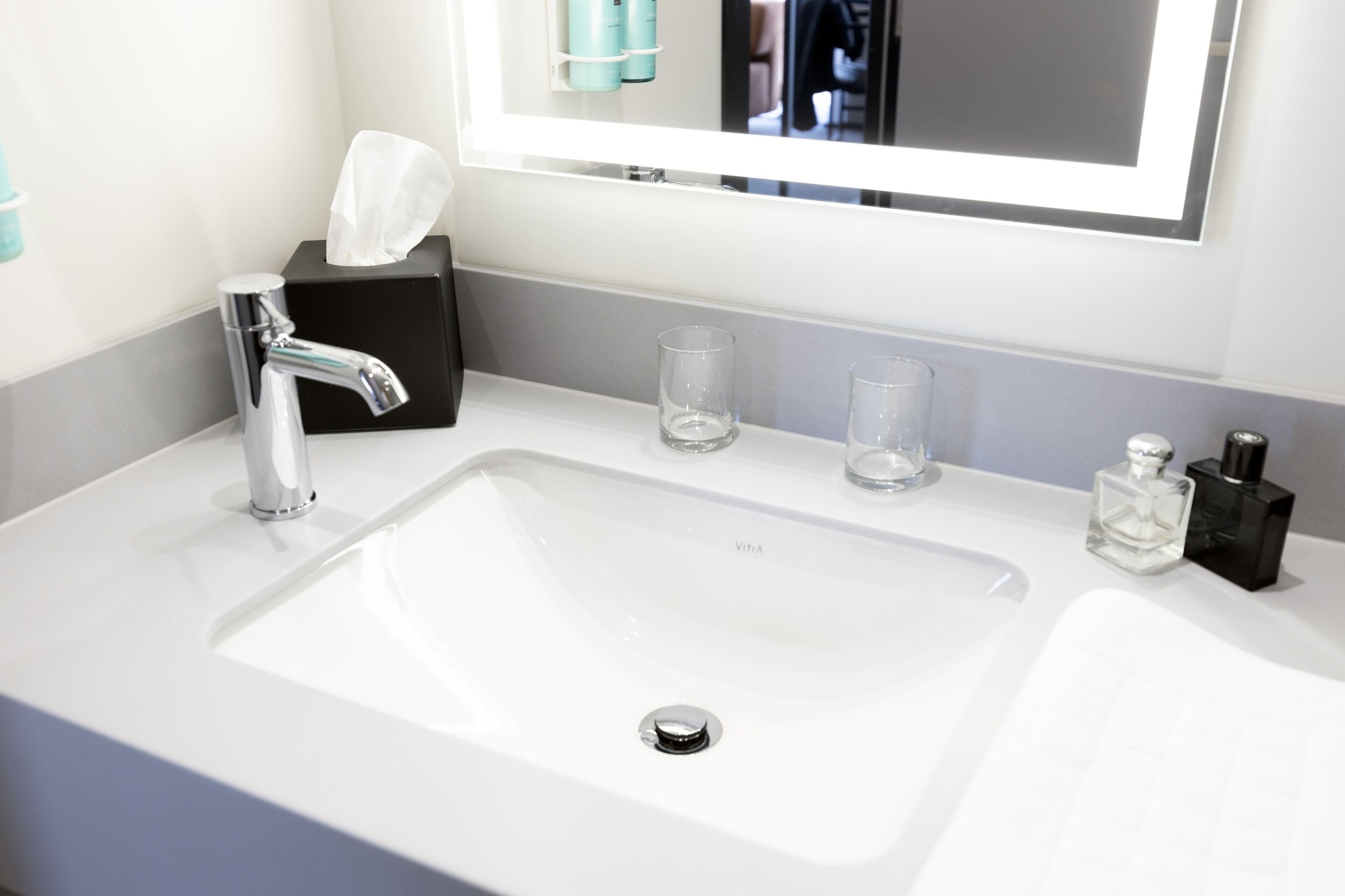 Un baño moderno con un lavamanos blanco, un espejo, una caja de pañuelos, dos vasos y una botella de perfume.