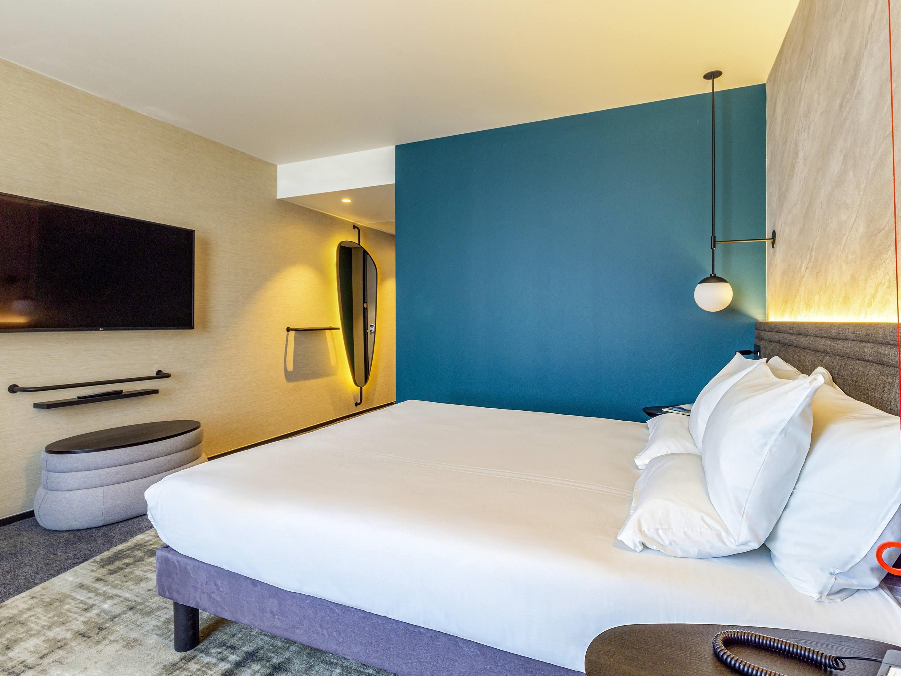 Quarto de hotel moderno com uma cama grande, uma televisão de tela plana na parede, uma mesinha de cabeceira e um banquinho redondo pequeno.