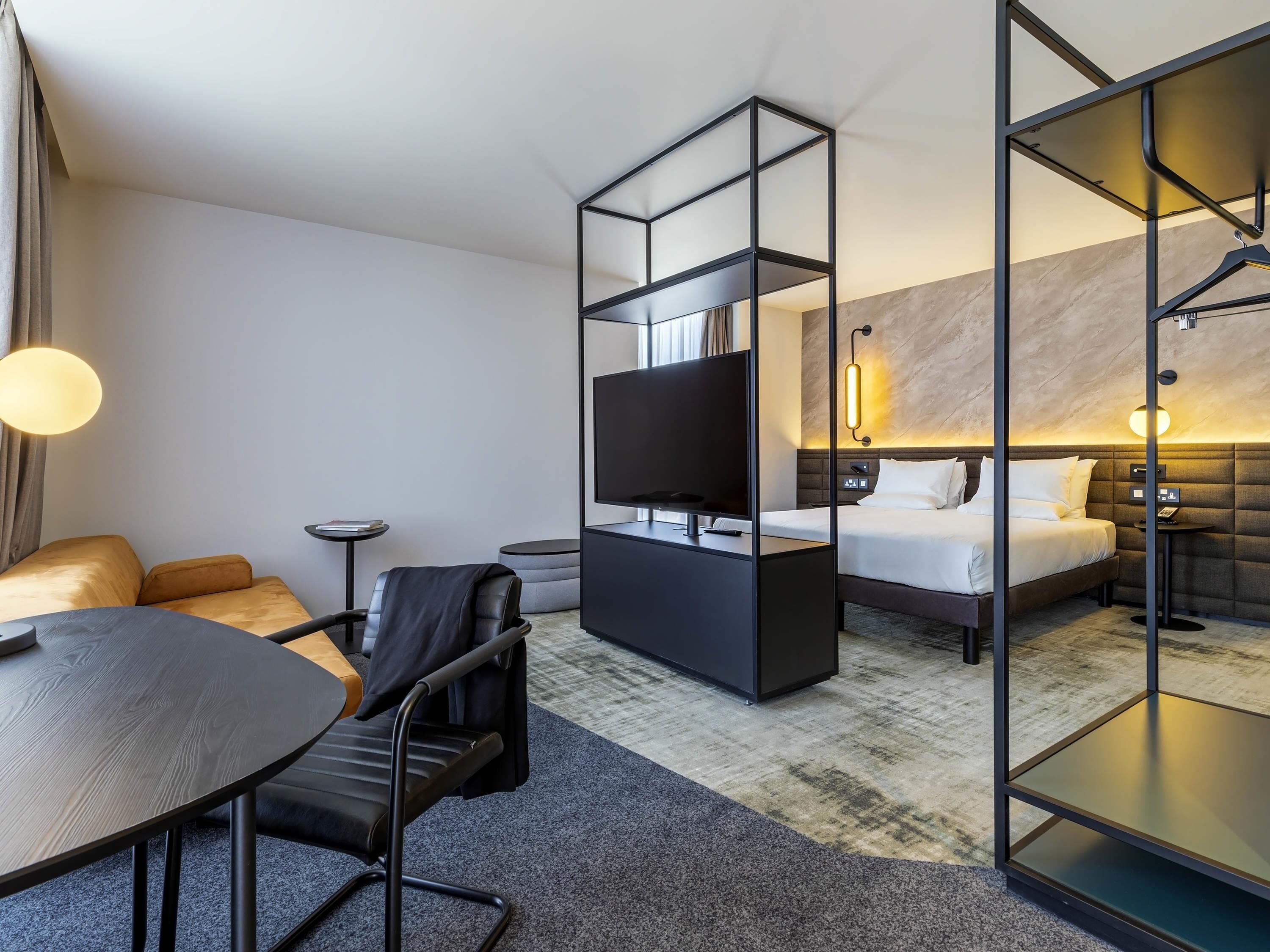 A modern hotel room with a bed, a desk, a chair, a TV, and a glass partition.