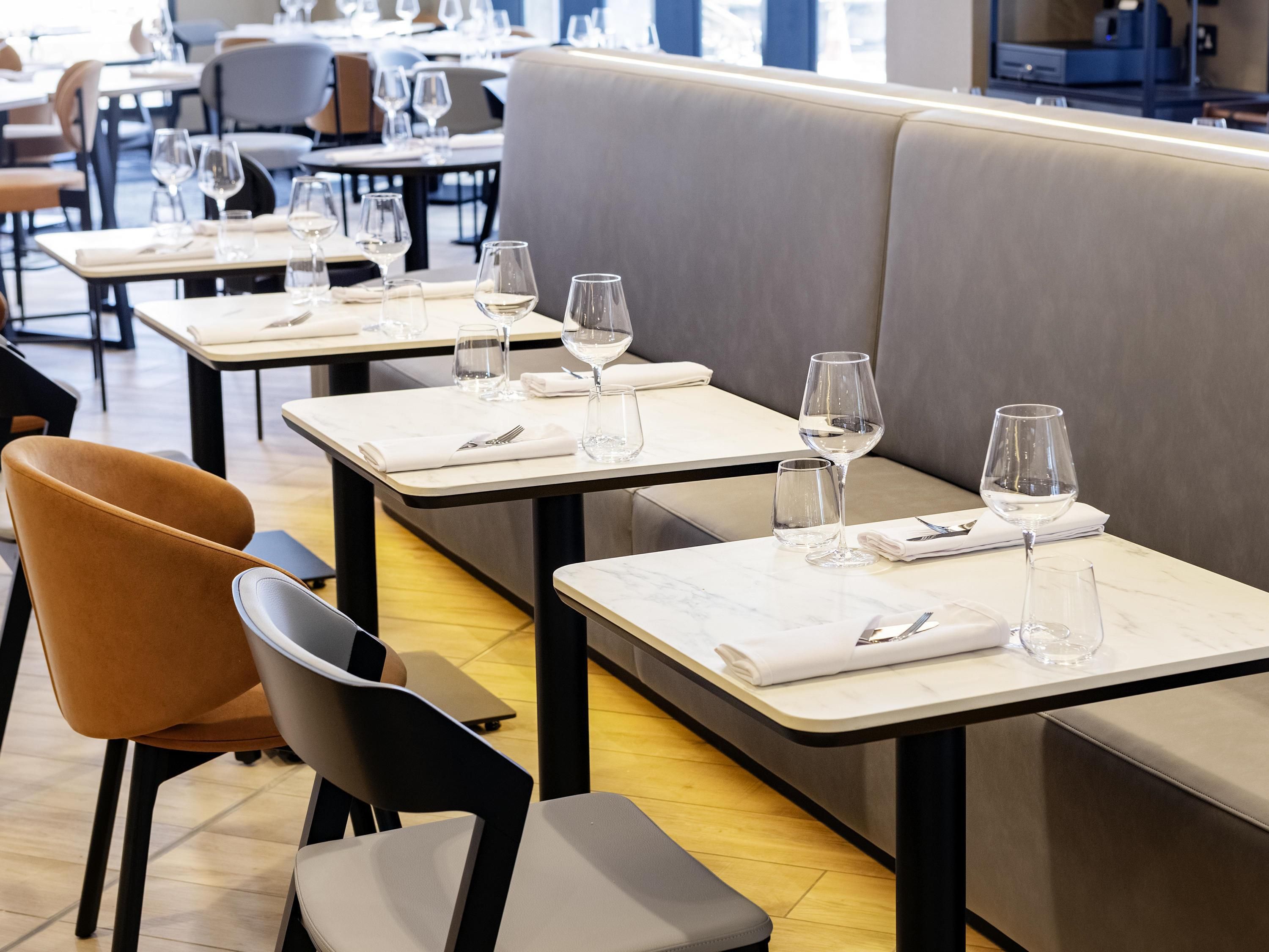 A restaurant interior with multiple tables and chairs, each set with a white tablecloth, wine glasses, and cutlery.