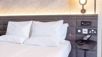 A hotel room with a bed, a bedside table with a phone, and a wall-mounted control panel.