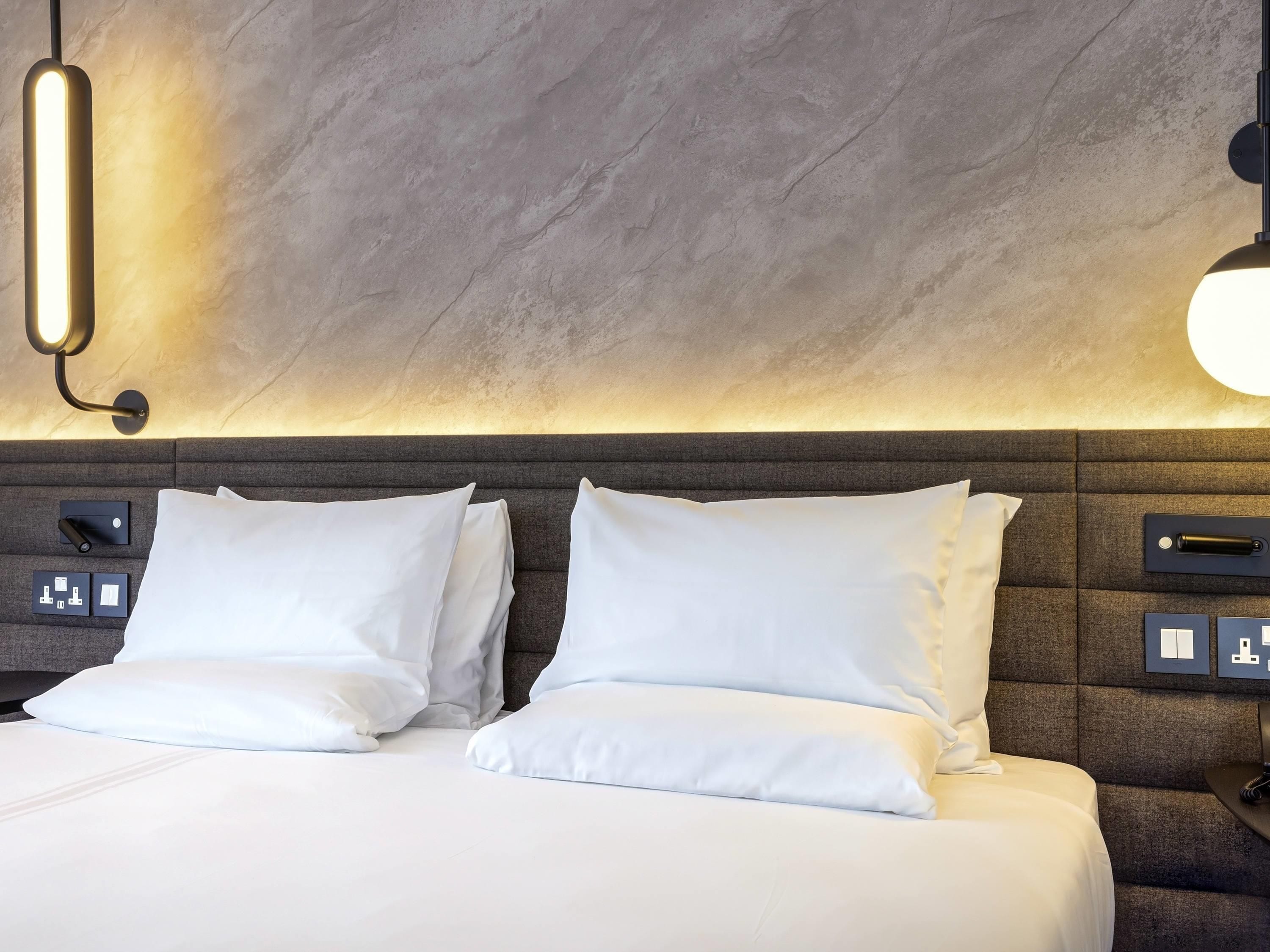 A hotel room with a bed, two pillows, a headboard with controls, and a wall-mounted light fixture.