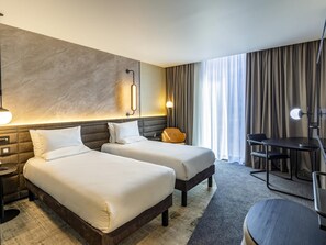A hotel room with two beds, a desk, a chair, a lamp, and a large window with curtains. - Novotel Liverpool Paddington Village (Liverpool)