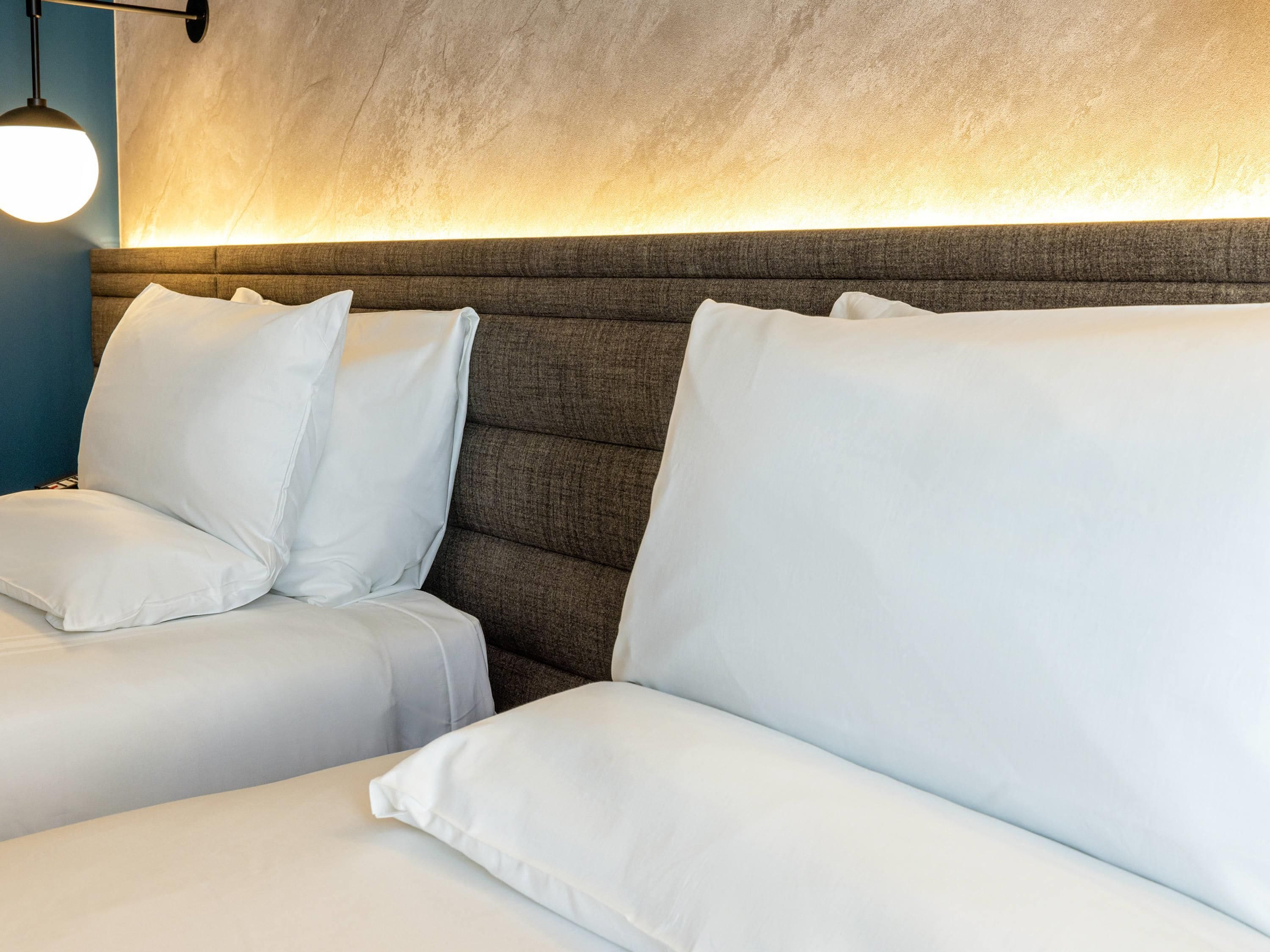 Una habitación de hotel con un cabecero grande y acolchado, y dos camas blancas con almohadas a juego.
