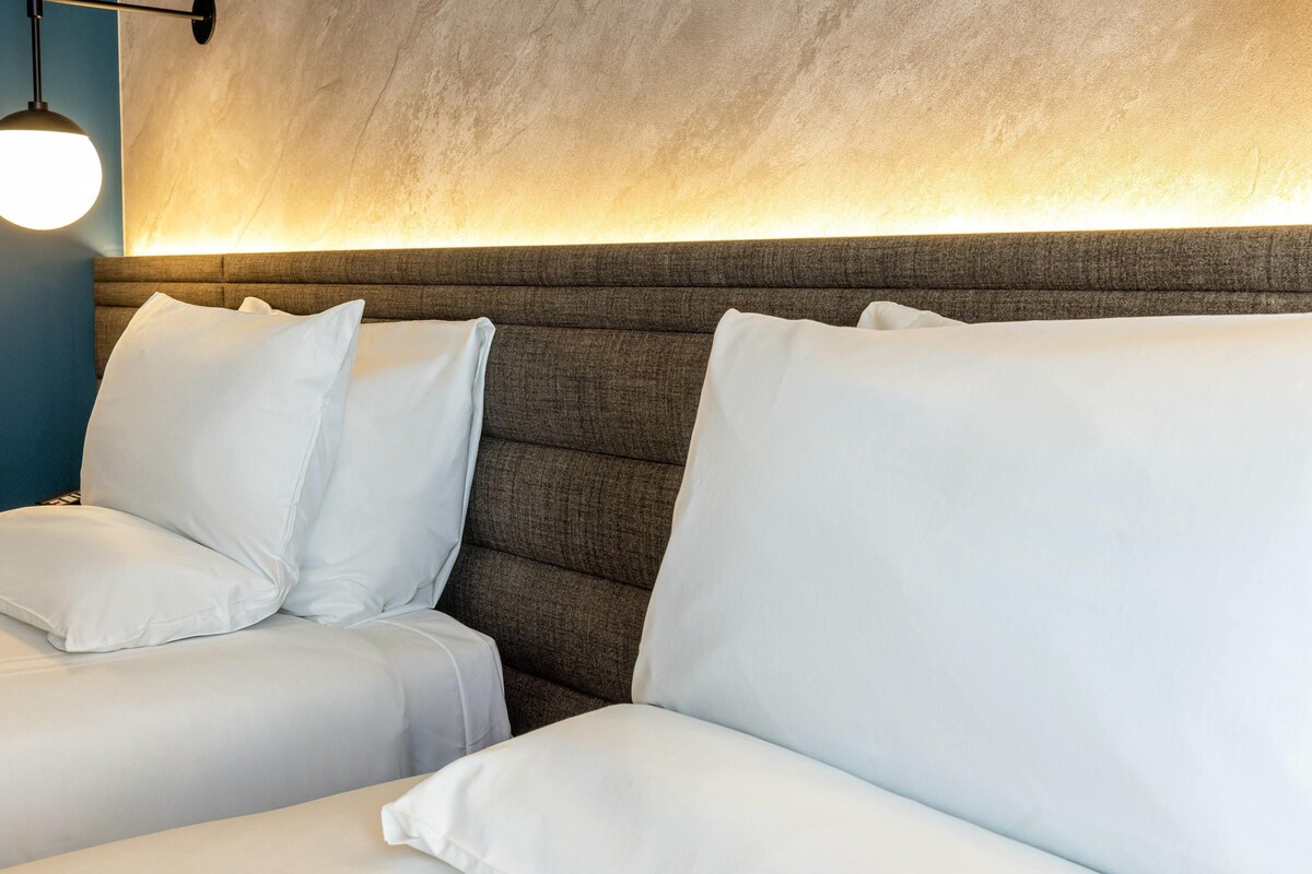 A hotel room with a large, padded headboard and two white beds with matching pillows.
