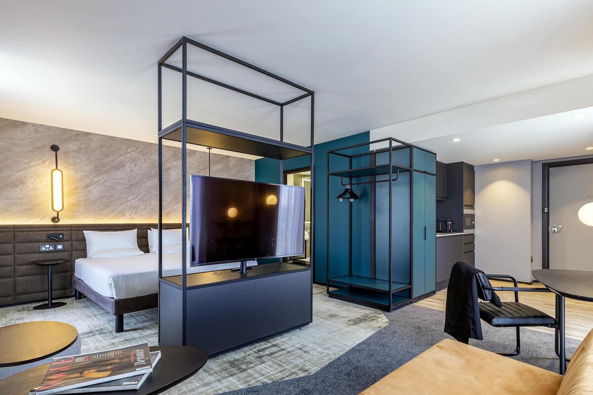 A modern hotel room with a bed, a TV, a seating area, and a wardrobe.