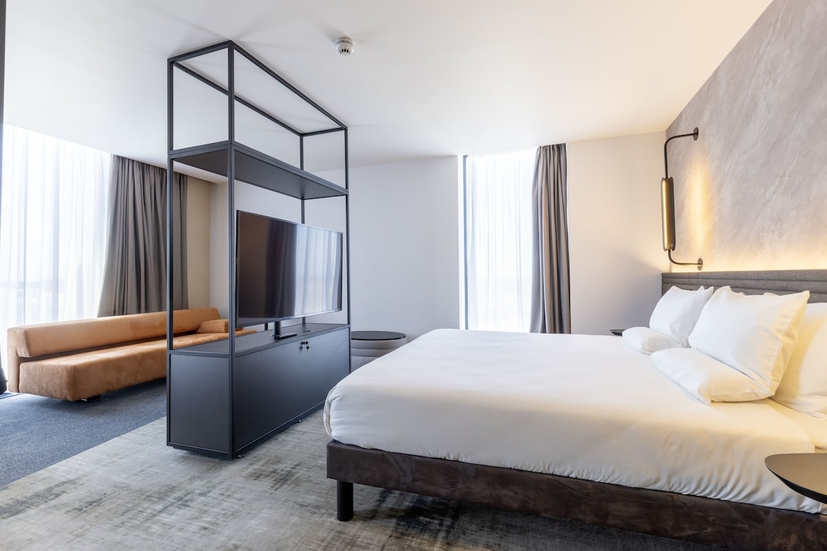 A modern hotel room with a large bed, a sofa, a TV, and a wardrobe.