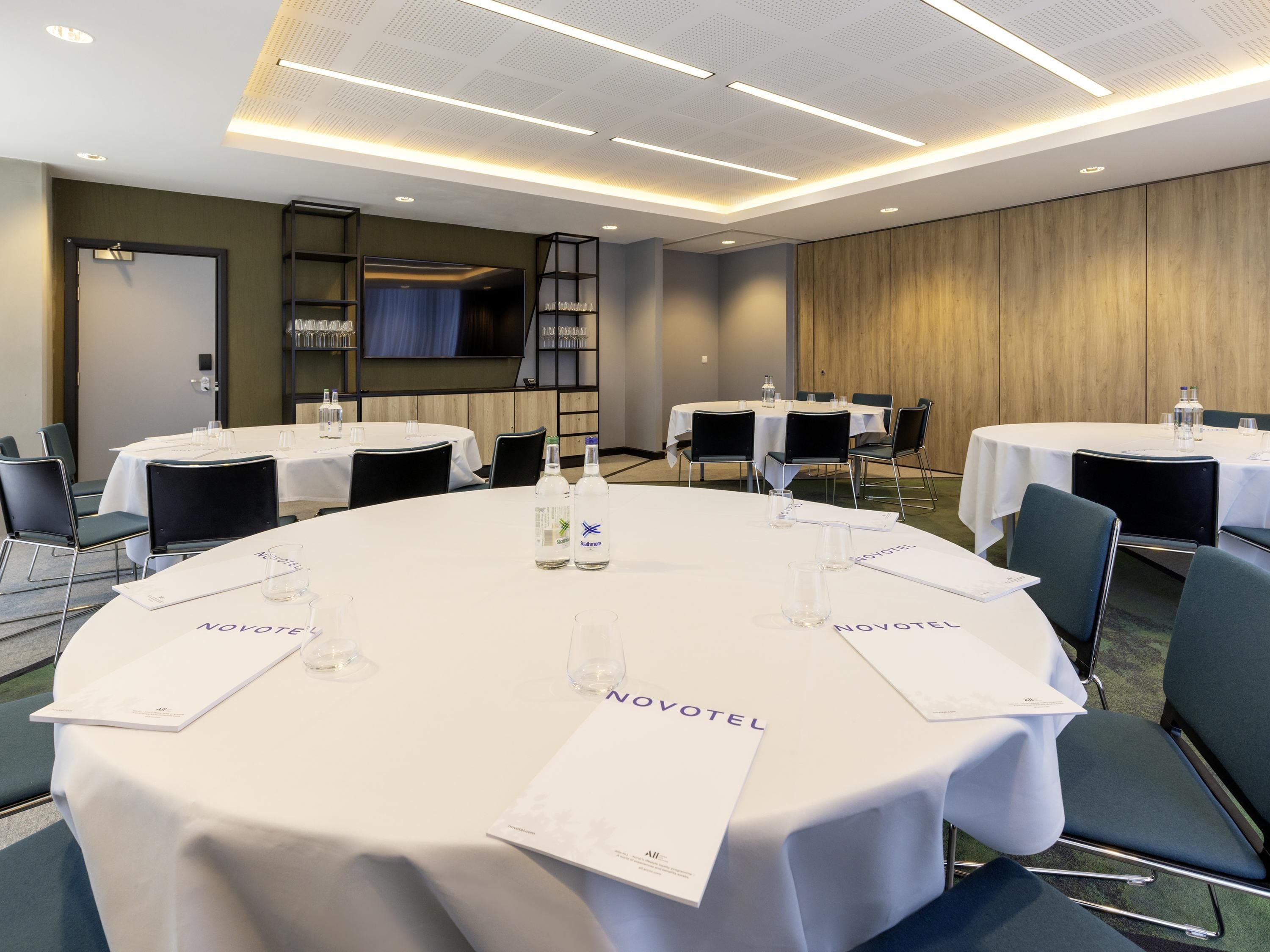 A conference room with round tables, each with a white tablecloth and name placard, equipped with water bottles and glasses.