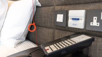 Una cama con un almohadón blanco, un objeto rojo colgando de la cama, un panel de control con un botón de RESTABLECER y un enchufe en la pared.