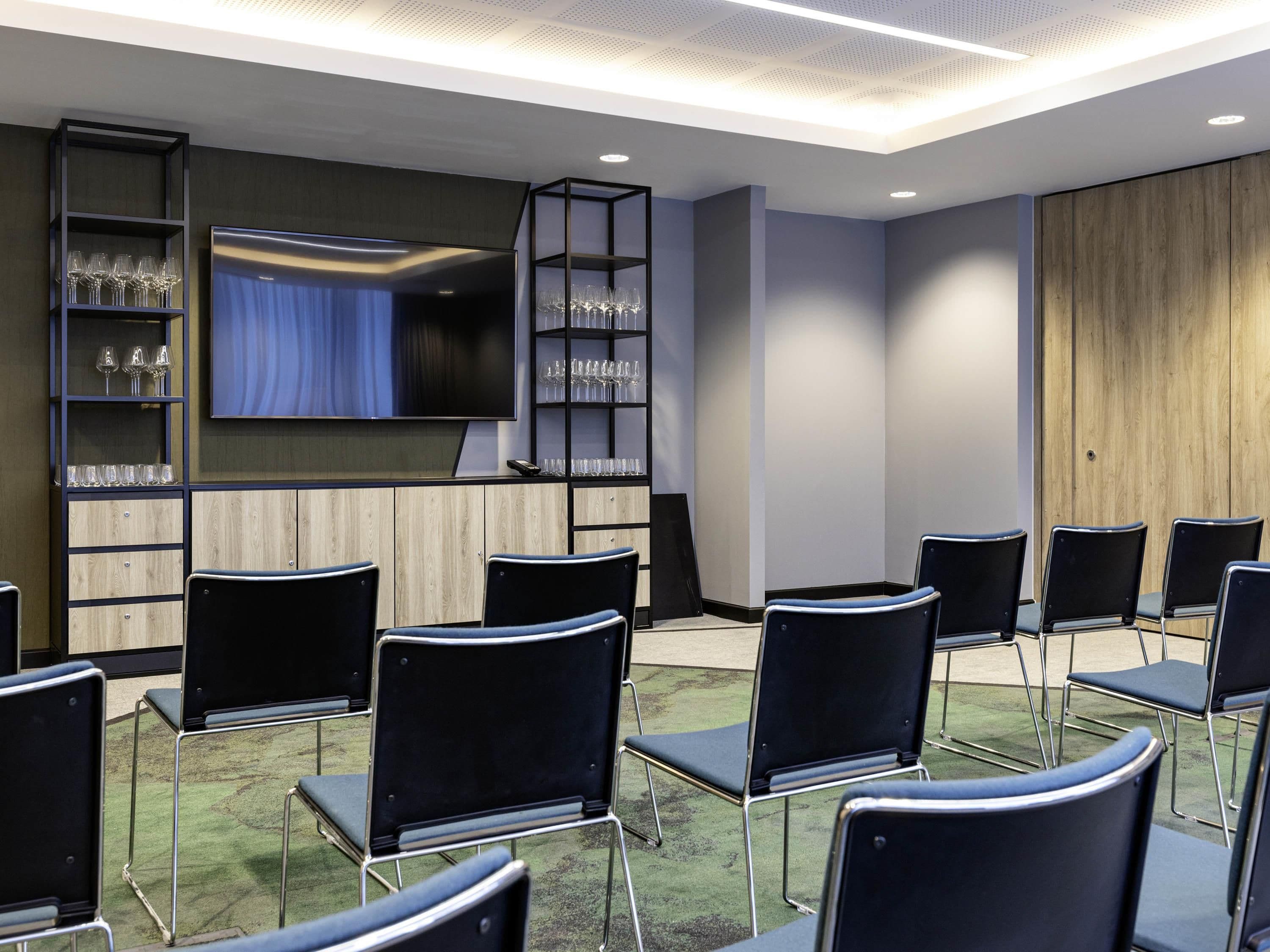 A conference room with a flat-screen TV, black chairs, and glassware on shelves.