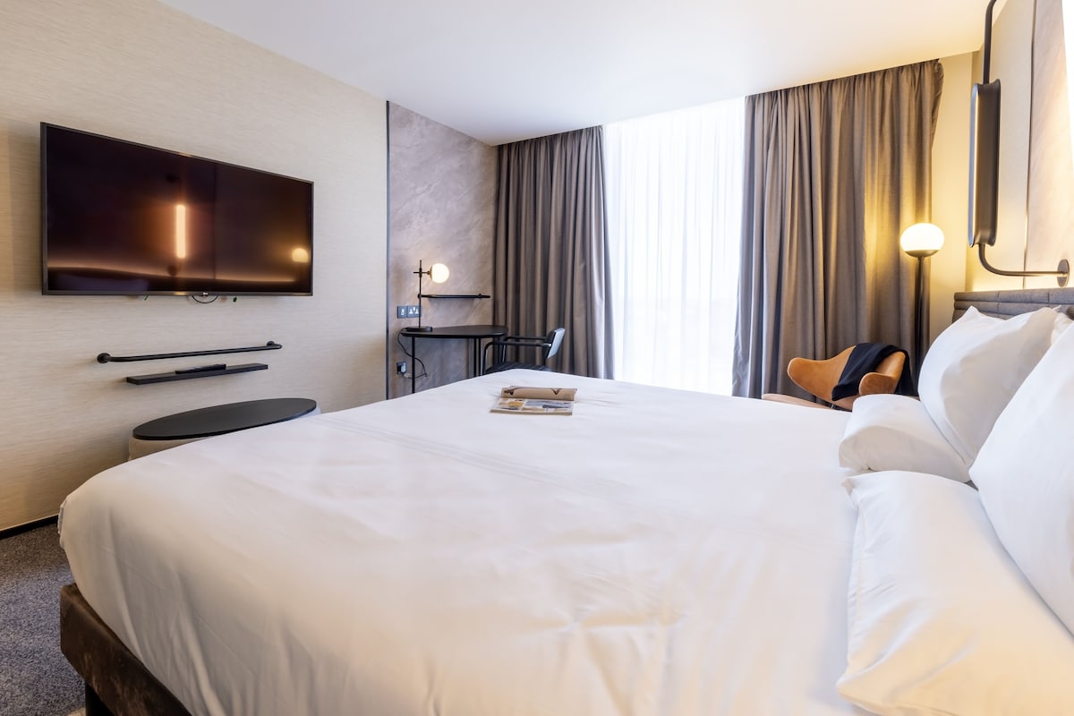 A hotel room with a large bed, a flat-screen TV mounted on the wall, a small desk with a chair, and a window with curtains.