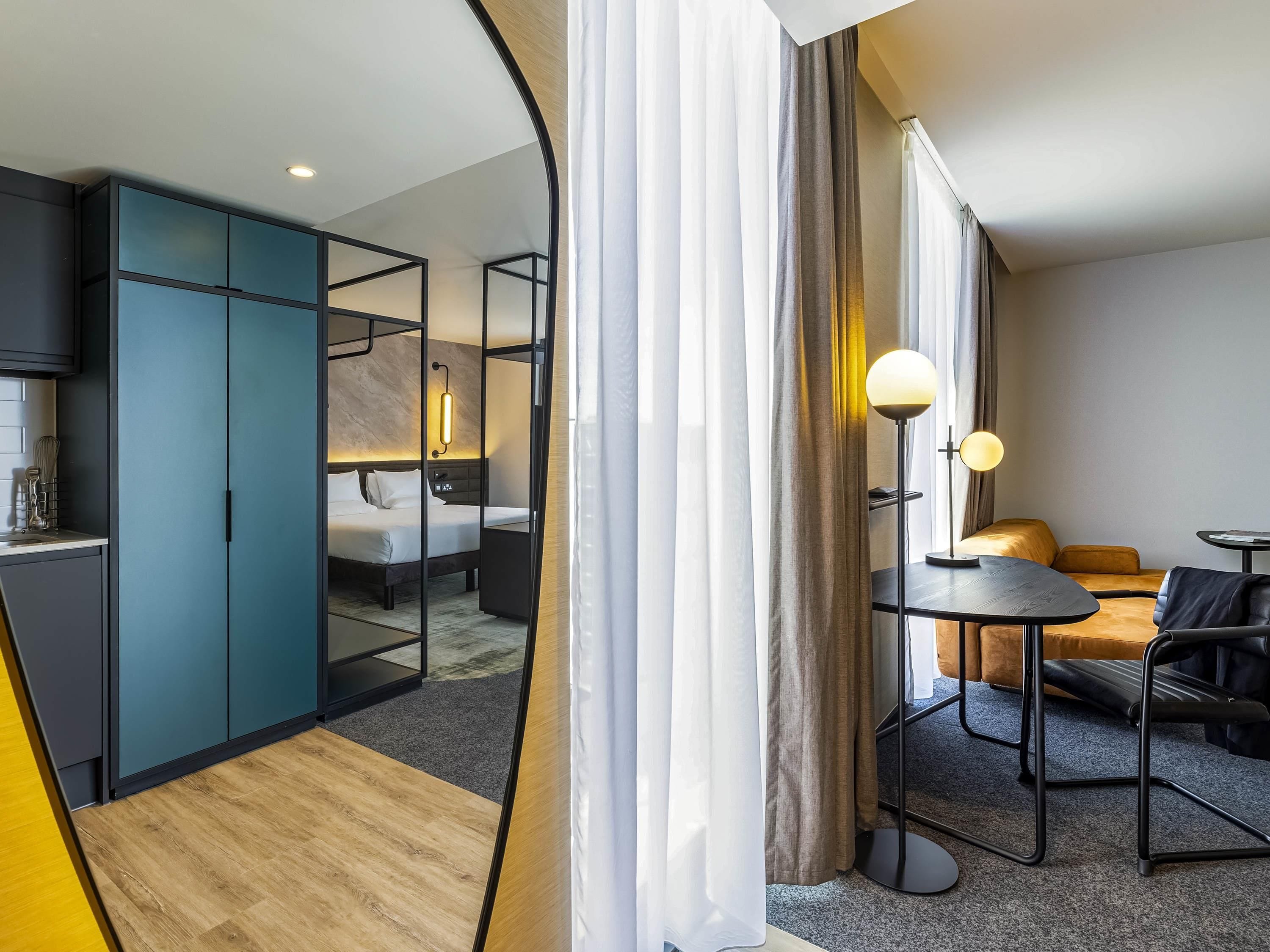 A modern hotel room with a bed, bedside table, lamp, and a seating area with a table and chairs.