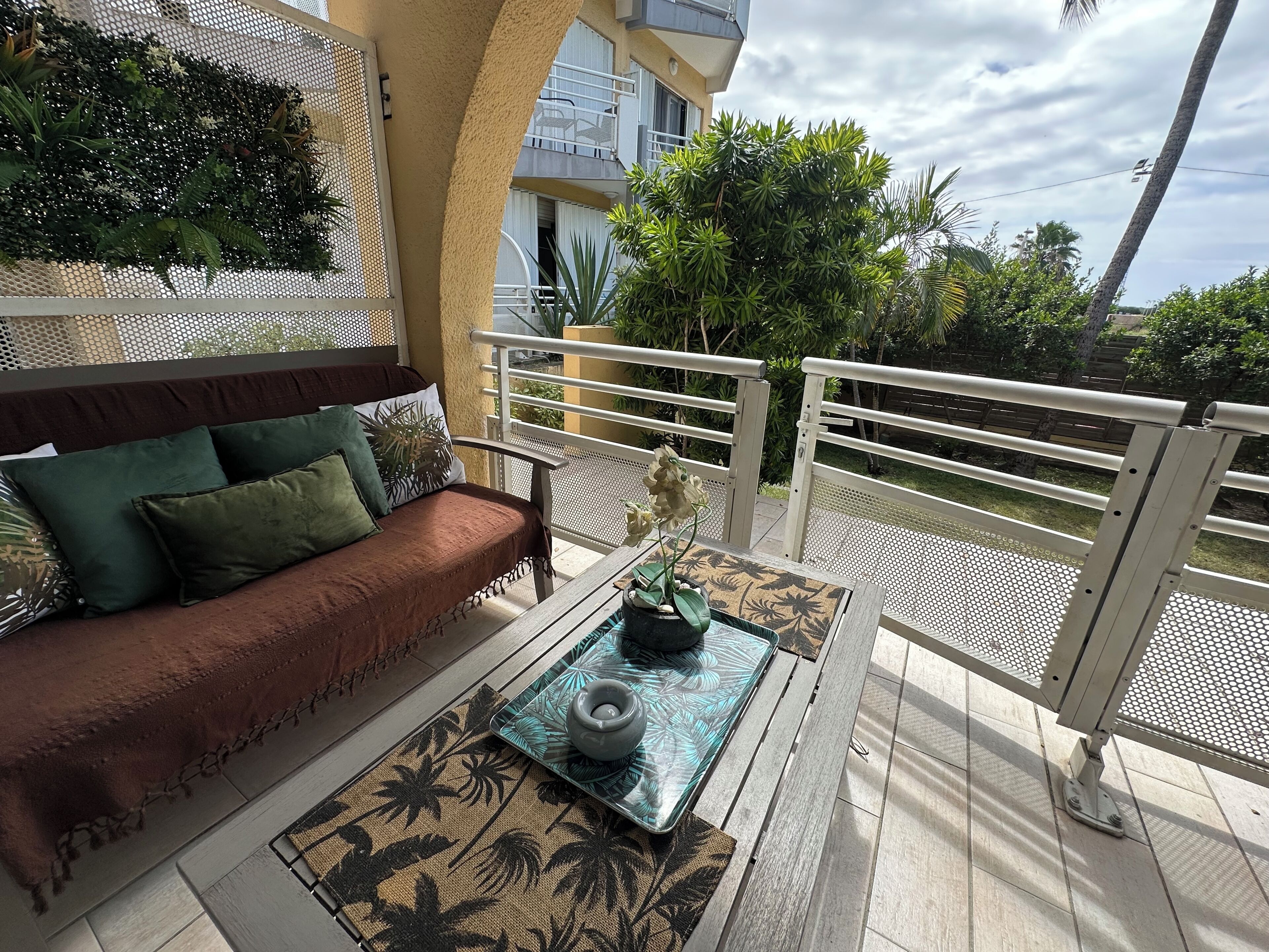 Ein Balkon mit einer Couch und einem Tisch, umgeben von einem weißen Geländer, mit Blick auf Grünflächen und einem Gebäude im Hintergrund.