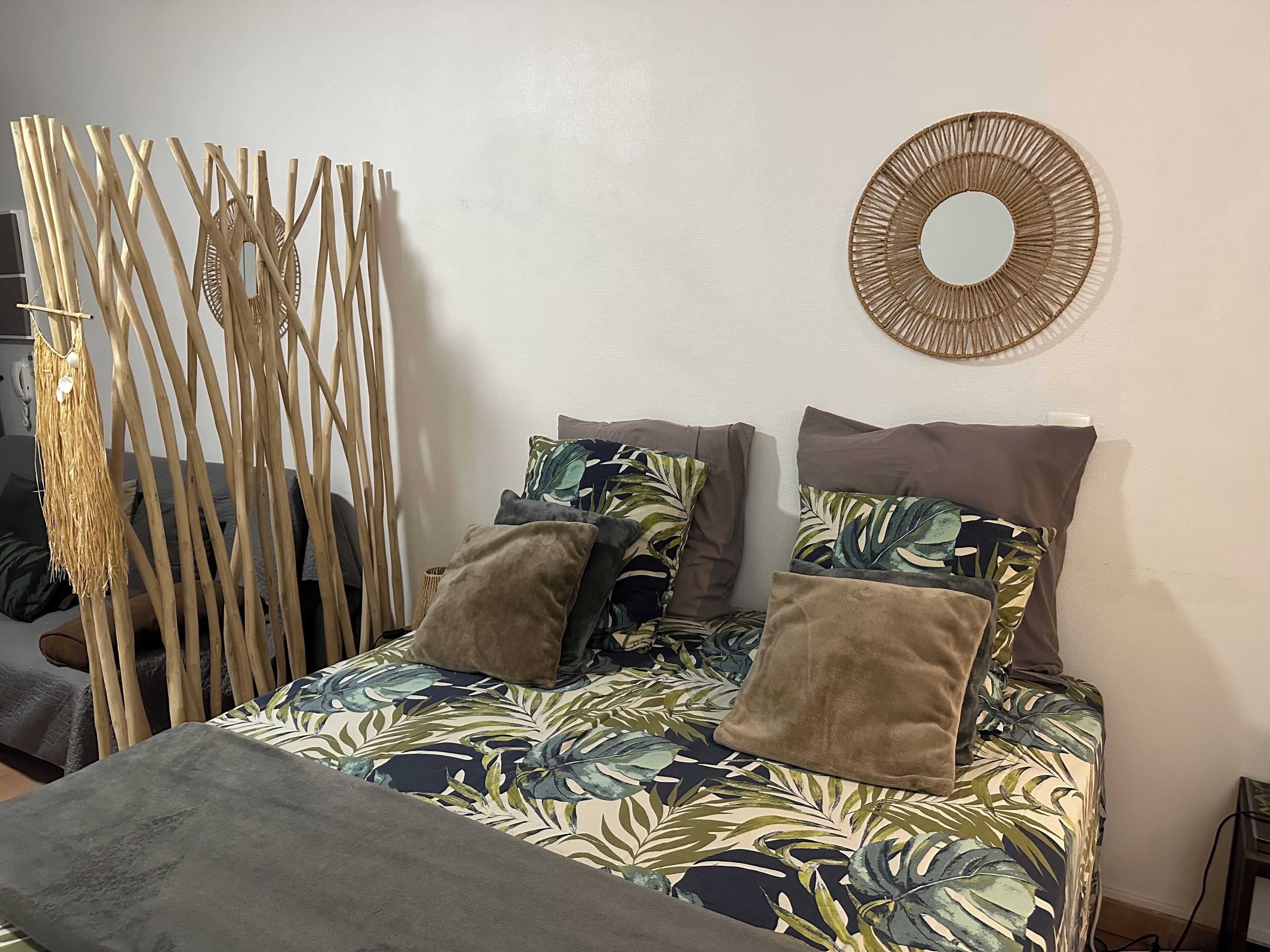 A bed with a tropical leaf-patterned bedspread, two brown pillows, and a woven wall decoration.