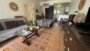 A living room with a sofa, coffee table, and a TV. There is a kitchen area with a dining table and chairs. - Studio "Entre Terre et Mer" Residence Anse des Sables (Marigot)