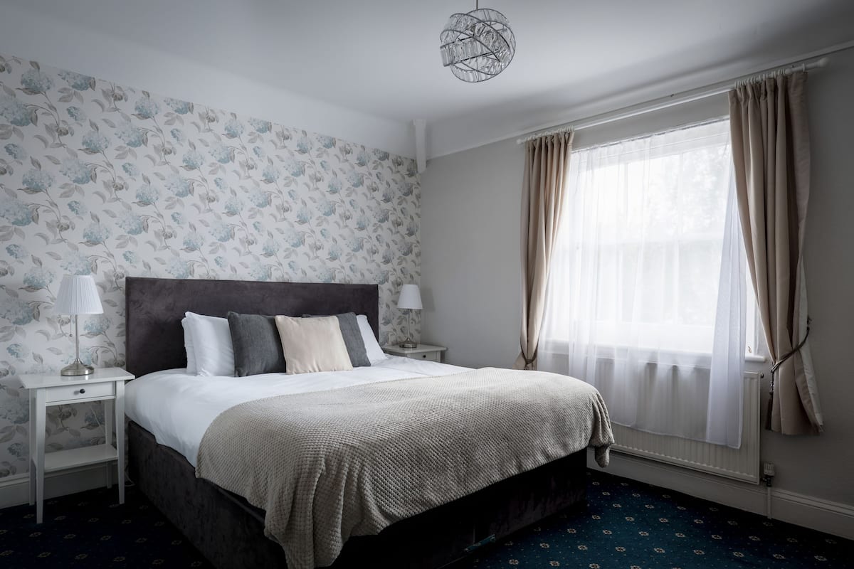 Een slaapkamer met een bed, nachtkastjes, een lamp, een raam met gordijnen en behangpapier met een patroon.