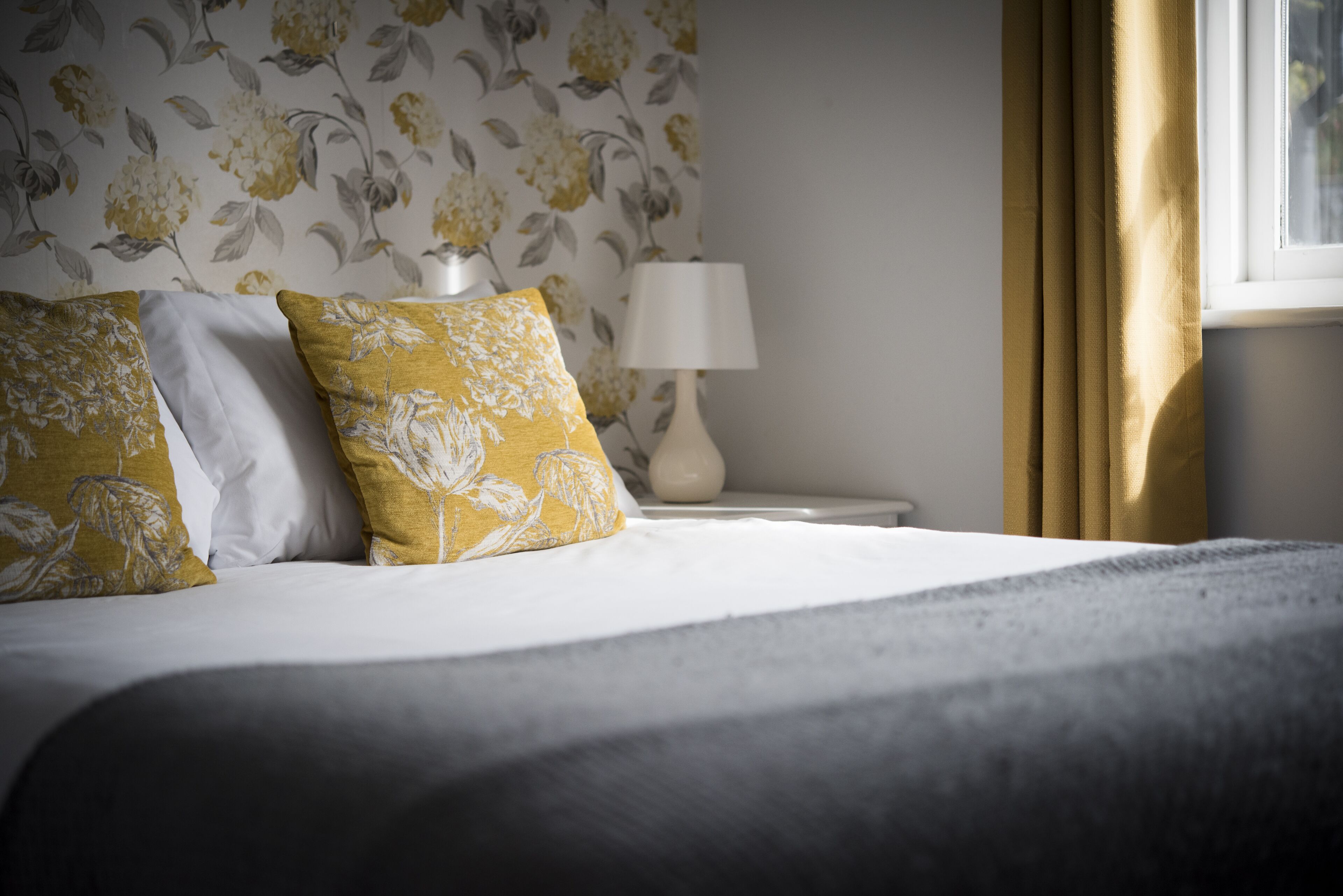 A neatly made bed with floral-patterned pillows, a bedside lamp, and curtains on a window.
