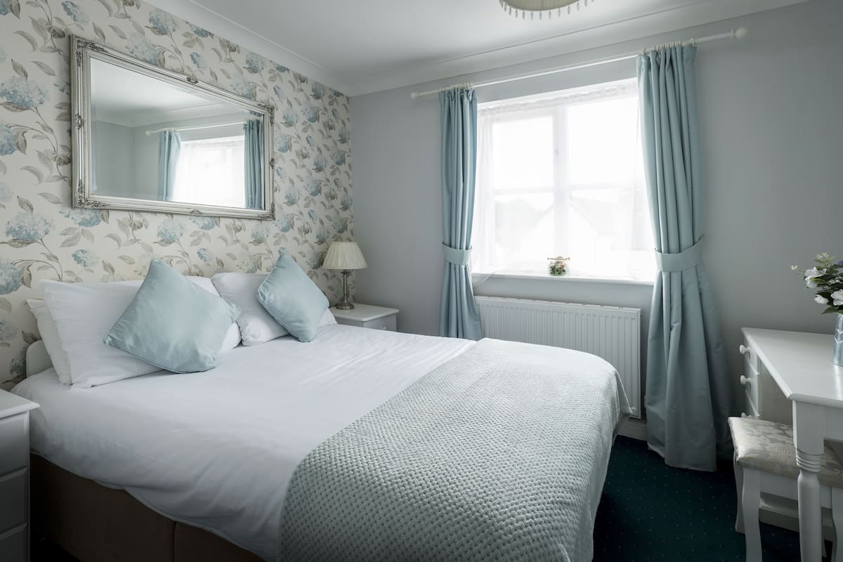 Een netjes opgemaakt slaapkamer met een groot bed, een nachtkastje met een lamp, een dressoir en een raam met gordijnen.