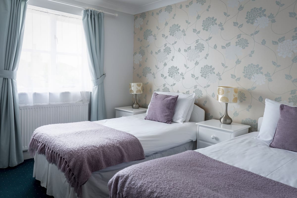 Een hotelkamer met twee bedden, een raam met gordijnen, bloemenbehang en bedlampen.
