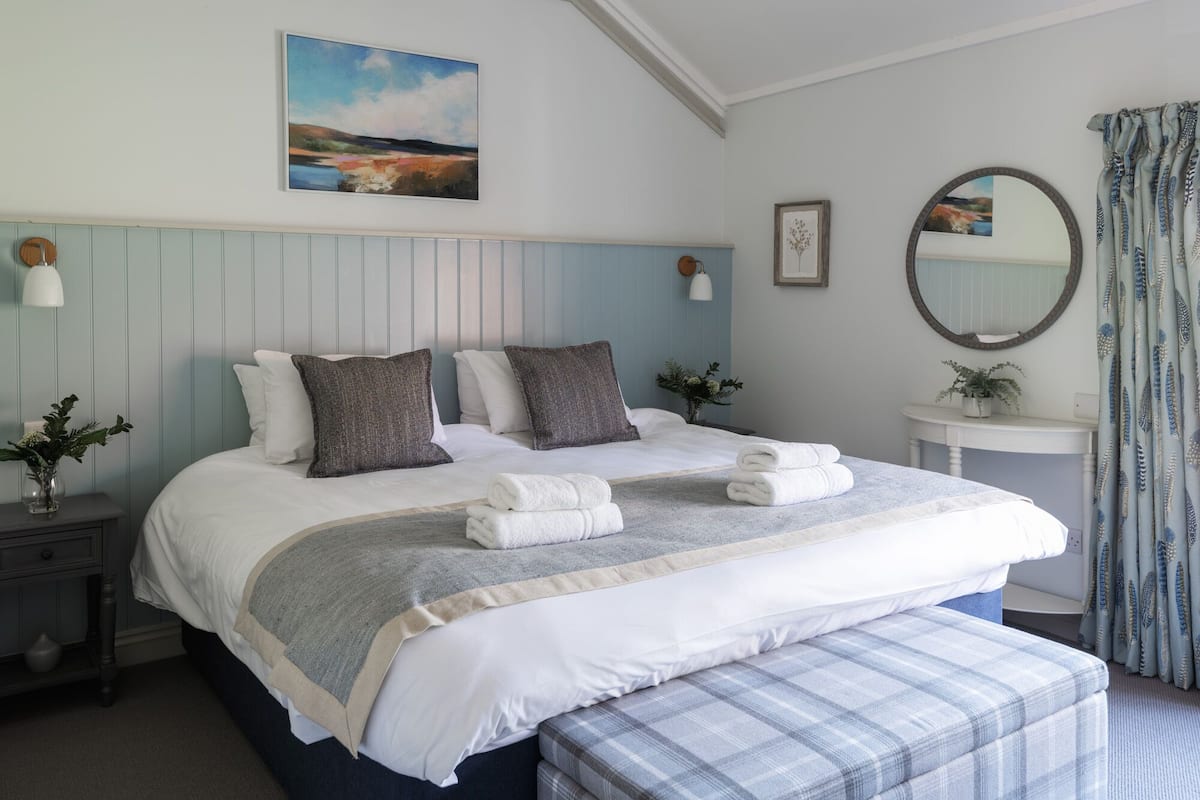 Een keurig opgemaakt slaapkamer met een bed, nachtkastjes, een bedlampje, een spiegel en een raam met gordijnen.
